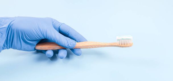 Ένα χέρι γάντια κρατά μια οδοντόβουρτσα μπαμπού με πάστα στα αριστερά σε ένα μαλακό μπλε φόντο, close-up άποψη πλευρά. - Φωτογραφία, εικόνα