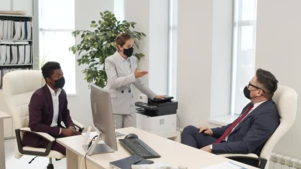 Colpo medio di dirigente maschile in maschera facciale e dei suoi colleghi che discutono nel suo ufficio mentre lavorano in mezzo a una pandemia - Filmati, video