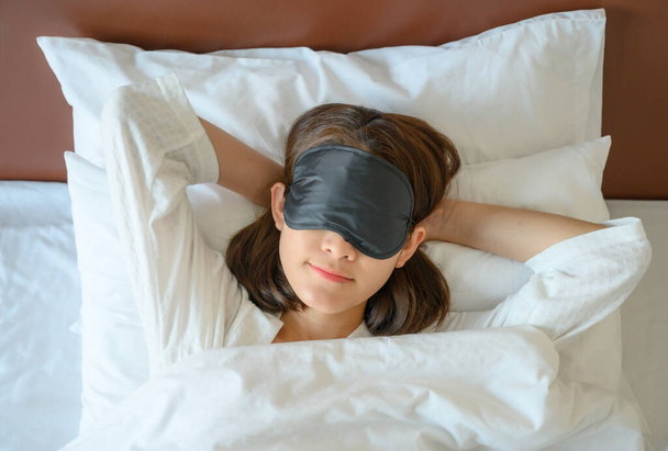 ベッドの上で眠っている間に睡眠マスクを身に着けている若い美しい女性の高い角度ビュー。睡眠マスクは、睡眠時に光を遮断することができ、ユーザーがより深いレベルの睡眠を達成することができます. - 写真・画像