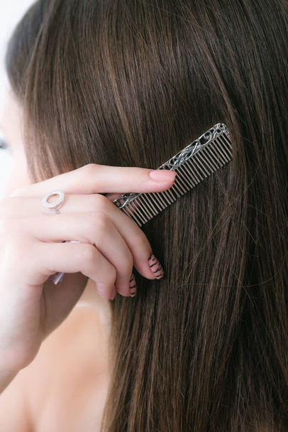 An attractive young girl with long dark hair uses a silver comb to style her hair. Beauty and fashion  - Φωτογραφία, εικόνα