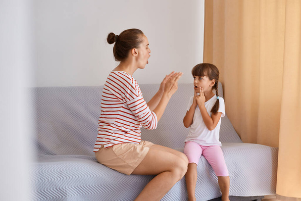 Image of professional female speech pathologist having lesson with little girl, people sitting on comfortable sofa near window, improving speaking, practicing sounds pronunciation. - Foto, Imagen
