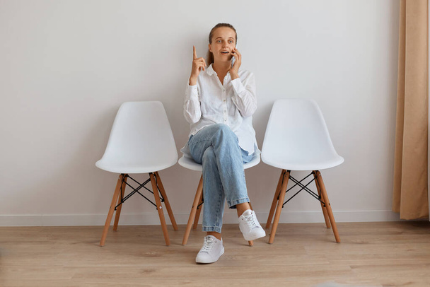 Indoor shot of attractive female with ponytail sitting on chair against light wall, woman wearing stylish jeans and white shirt, talking cell phone, having idea and pointing finger up. - Foto, Imagem