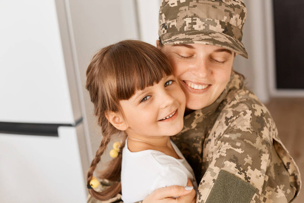 Indoor shot of happy military woman wearing camouflage uniform returning home after army, meeting her lovely daughter, hugging her child with great love. - Foto, afbeelding