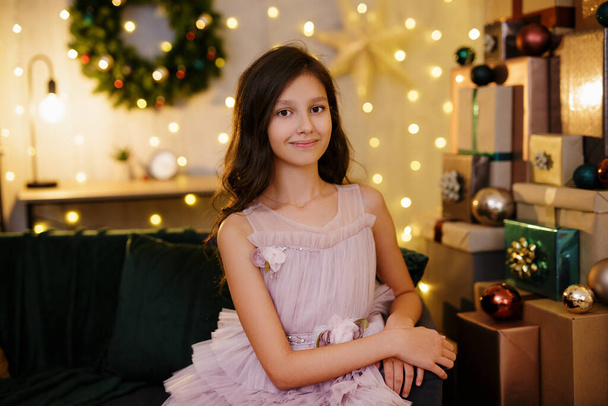 bonito preteen menina no vestido festivo sentado no quarto decorado com luzes led e montão de presentes de Natal - Foto, Imagem
