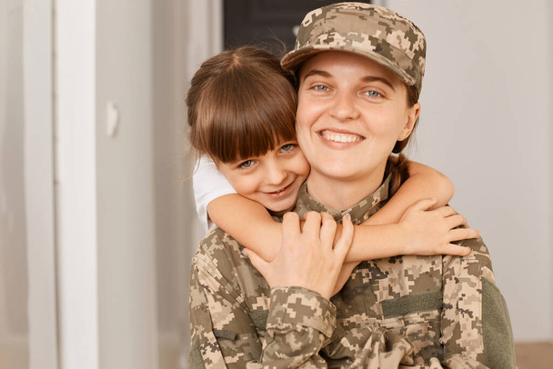 Portrait of female with toothy smile posing with her daughter after returning from army or exercising, kid embracing her mommy, missing her little girl. - Valokuva, kuva
