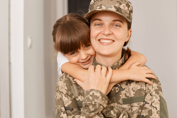 Πορτρέτο της ευτυχισμένης οικογένειας ποζάρουν εσωτερικά μετά την επιστροφή της μητέρας στο σπίτι μετά το στρατό, μικρό θηλυκό παιδί αγκαλιάζει τη μητέρα της, οι άνθρωποι κοιτάζοντας κάμερα με θετική έκφραση. - Φωτογραφία, εικόνα