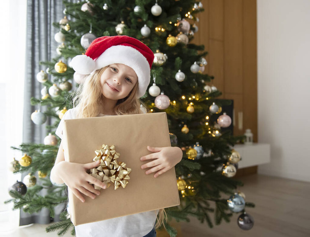 Een meisje met cadeaus speelt bij de kerstboom. Woonkamer interieur met kerstboom en decoraties. Nieuwjaar. Cadeau geven. - Foto, afbeelding