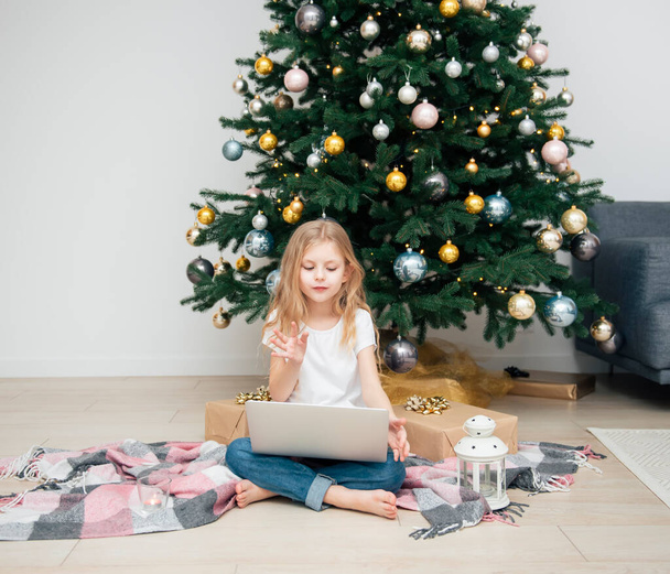 Vánoční stromek ve velkém obývacím pokoji. Holčička si hraje u vánočního stromečku. Dívka s laptopem komunikuje s přáteli. - Fotografie, Obrázek