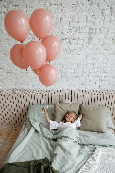 Το κορίτσι χαμογελάει χαρούμενα και κρατάει ροζ μπαλόνια, ξαπλωμένη στο κρεβάτι με μαξιλάρια. Κομψό εσωτερικό, ευτυχισμένη παιδική ηλικία, εορτασμός γενεθλίων. - Φωτογραφία, εικόνα