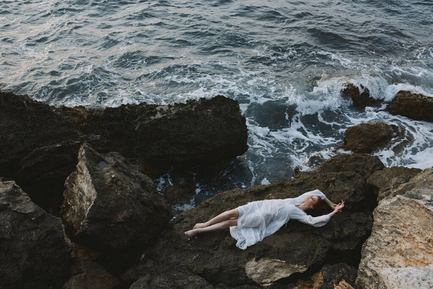 Mezítlábas nő egy félreeső helyen egy vad sziklás parton, fehér ruha kilátással felülről - Fotó, kép