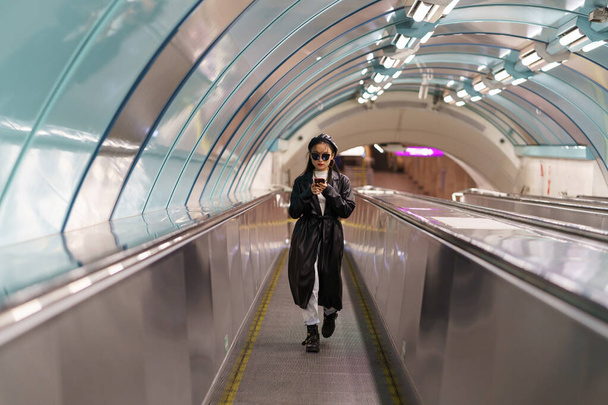 Blogger chica japonesa en trinchera de moda y gafas en el uso de escaleras mecánicas smartphone leer el comentario seguidor - Foto, Imagen