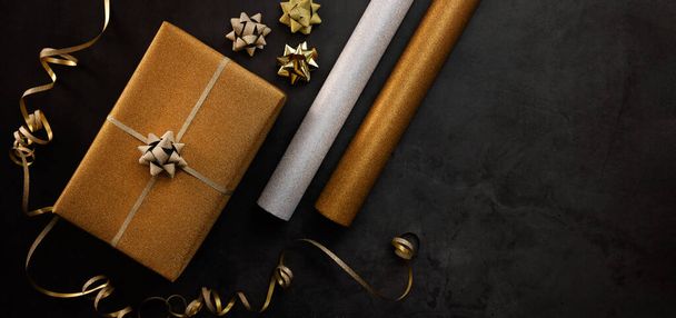 gift wrapping service. wrapped golden present box with wrapping paper rolls on dark stone background. banner copy space - Фото, изображение