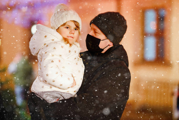 Kisiskolás lány ül az apa vállán a karácsonyi piacon Németországban. Boldog kisbabát és férfit orvosi maszkkal a koronavírus világjárvány miatt. Család, kötődés, szerelem. Családi karácsony ideje. - Fotó, kép