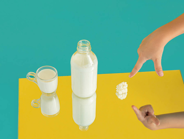 Botella, taza de leche de kéfir y mano de niño señala hongos de kéfir. Mínimo lácteo natural saludable comida escena reflejada en el espejo sobre fondo azul amarillo. Idea de origen de bebidas digestivas o probióticos.  - Foto, Imagen