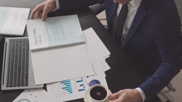 Blick von oben auf einen männlichen CEO in blauer Festkleidung, der im Büro am Schreibtisch sitzt, Unternehmensdokumente liest und schwarzen Kaffee aus der Tasse trinkt - Filmmaterial, Video