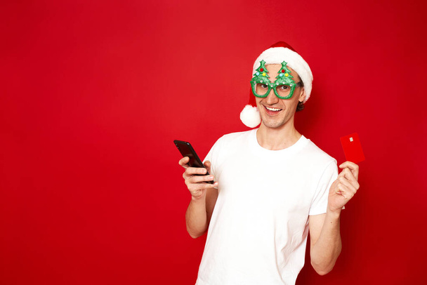 concept of people, online shopping, technology, discounts. an excited man in glasses Christmas trees Santa Claus hat makes an order via phone pays credit bank card, isolated on red studio background - Foto, afbeelding