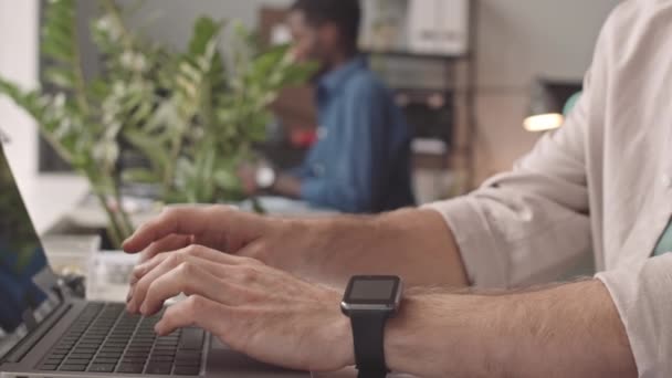 Low angle close-up of hands of male employee wearing shirt and smartwatch sitting at desk in office at daytime, typing on portable computer - Footage, Video