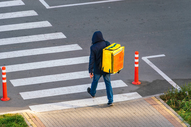 Un mensajero que entrega mercancías con una gran bolsa amarilla en su espalda camina a lo largo del cruce de cebra en el cruce peatonal a la luz del sol. Entrega de mercancías de los almacenes durante la pandemia del coronavirus - Foto, imagen