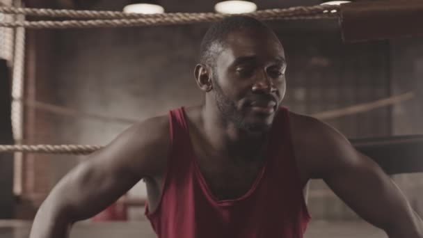 Közepes közelkép lassú portré izmos afro-amerikai férfi bokszoló piros mez és boksz kesztyű nézi a kamera és dob puncs, ül a boksz gyűrű - Felvétel, videó