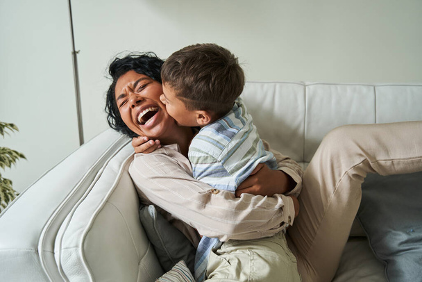 Cute little boy embracing his lovely mother while sitting at the sofa - Foto, Imagen