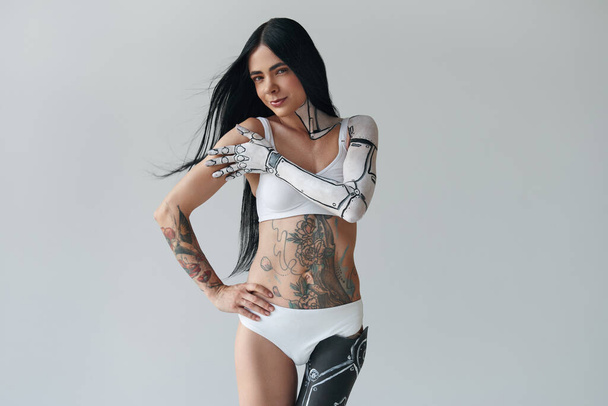 Horizontal portrait of a brunette tattooed girl with cyborg body art and prosthesis leg. Woman looking at the camera while posing at the studio. Unusual appearance concept - Zdjęcie, obraz