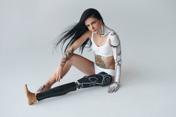 Unusual appearance. Full length view of the woman with prothesis leg and cyber body art sitting at the floor and posing at the studio. People with special needs concept - 写真・画像