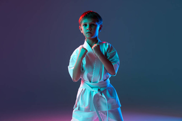 Retrato de niño pequeño en kimono blanco entrenando karate aislado sobre fondo azul degradado en neón. Puños arriba - Foto, imagen