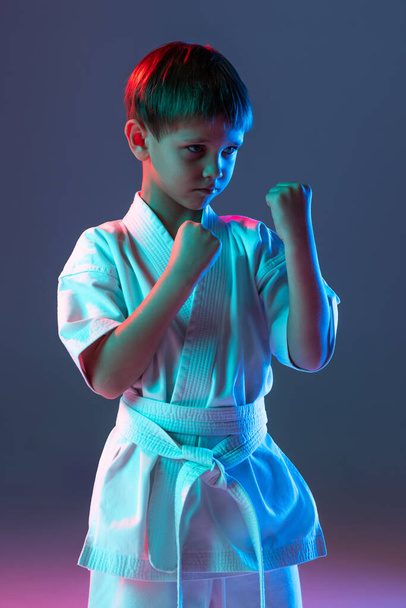 Cropped portrait of child, concentrated sportsman training, practising karate isolated over gradient blue background in neon - Foto, afbeelding