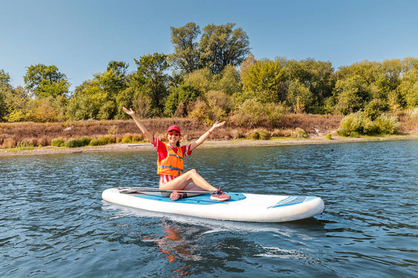 Happy woman wearing life vest sitting on an inflatable sup board. Healthy recreation and leisure on a lake and water sports - Photo, Image