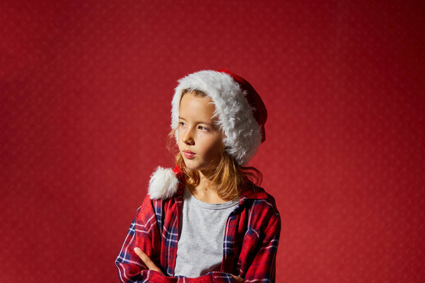 Рождественская маленькая девочка в Санта-Шляпе обиделся на кого-то, перепады настроения изолированные красный фон студии - Фото, изображение