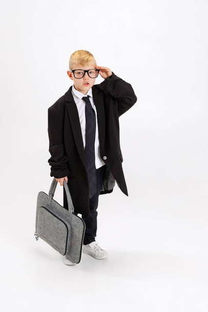 Close-up of cute little boy, kid businessman in huge jacket suit holding laptop case isolated on white studio background. - Foto, Imagem