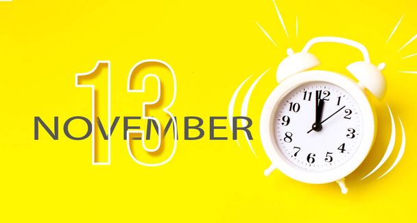 Il 13 novembre. Giorno 13 del mese, data del calendario. Sveglia bianca con giorno di calendario su sfondo giallo. Concetto minimalista di tempo, scadenza, orario di lavoro, mattina. Autunno mese, giorno dell'anno concetto - Foto, immagini