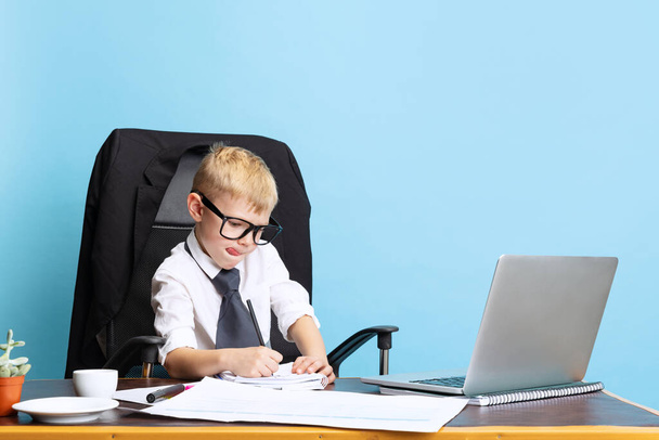 One little boy, child businessman sitting at office table isolated on blue studio background. Concept of smart childhood - Fotoğraf, Görsel
