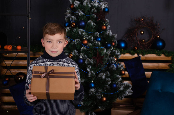 boldog jóképű csinos tinédzser szőke fiú kötött pulóverben gazdaság új év ajándék közelében egy karácsonyfa - Fotó, kép