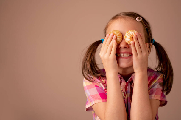 Funny portrait of young happy girl places the tangerine in her two eyes. Copy space.  - Zdjęcie, obraz