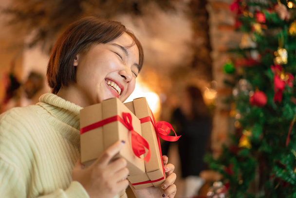 Happy beautiful woman in sweater is smiling and holding present box in front of the christmas tree and decoration for celebration christmas or new year in living room at home - Foto, Imagem