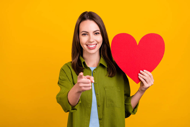 Photo of adorable pretty young lady wear green shirt holding large heart pointing finger you smiling isolated yellow color background - Valokuva, kuva