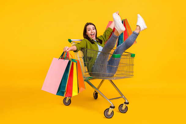 Photo of pretty funky young lady wear green shirt smiling holding shopping bags riding cart isolated yellow color background - Foto, afbeelding