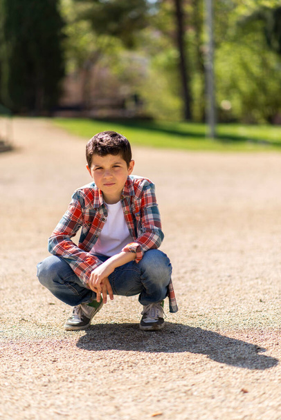 Χαρούμενο αγόρι που φοράει καθημερινά ρούχα ενώ ποζάρει στο πάρκο της πόλης. - Φωτογραφία, εικόνα