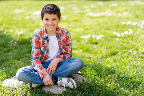 Χαρούμενο πορτρέτο αγόρι κάθεται στο γρασίδι του πάρκου της πόλης, ενώ κοιτάζοντας την κάμερα - Φωτογραφία, εικόνα