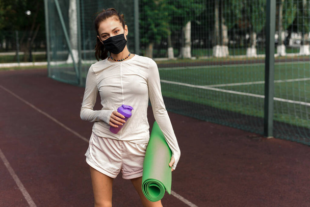 Une jeune femme portant un masque de protection noir se tient sur le terrain de sport, tenant un tapis de yoga et une bouteille d'eau. Activités sportives en quarantaine. À l'extérieur - Photo, image