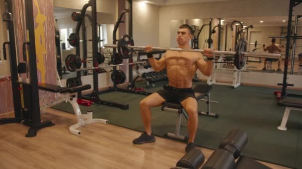 Um jovem fisiculturista treinando no ginásio e faz uma pressão de barra do peito sentado enquanto bombeia deltóides e tríceps - Filmagem, Vídeo