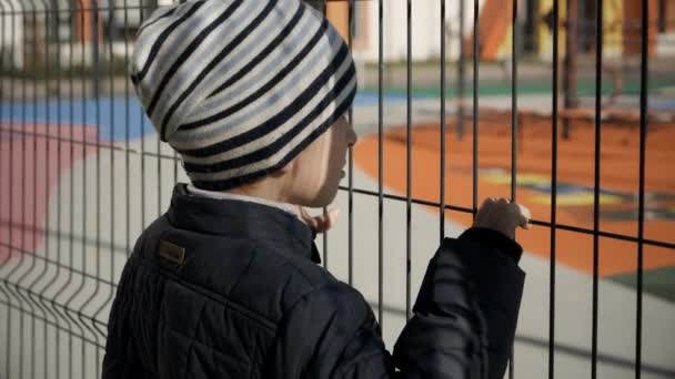 Sconvolto ragazzino stressato tenendo e guardando attraverso la recinzione di metallo alto sulla scuola costosa e parco giochi. Concetto di povertà, immigrazione, bullismo e stress infantile - Filmati, video