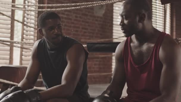 Medium slow shot di pugile afro-americano che ascolta il suo allenatore dimostrare pugni boxe seduti insieme sul ring durante l'allenamento - Filmati, video