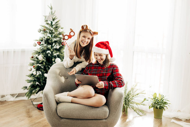 Dwie radosne siostry siedzą w domu obok choinki i gratulują przyjaciołom i dziadkom Bożego Narodzenia i Nowego Roku oraz wygłupiają się. Wirtualne pozdrowienia, gratulacje online - Zdjęcie, obraz