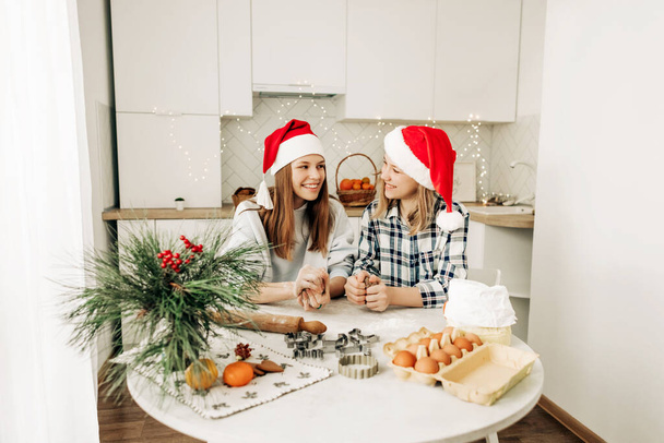 Two charming sisters in Santa Claus hats bake cookies for Christmas in the kitchen. Christmas cooking, preparation for the holiday, Christmas cookies - Photo, Image