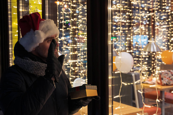 Hombre en sombrero de Santas mirando y soñando en escaparate iluminado - Foto, Imagen