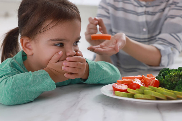 Mother feeding her daughter indoors, closeup. Little girl refusing to eat vegetables - Фото, изображение
