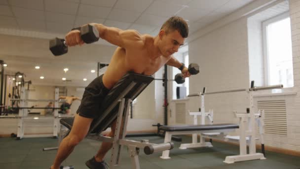 Młody muskularny mężczyzna z nagim tułowiem ćwiczy na siłowni i podnosi ramię z hantlami leżącymi na pochyłej ławce zwróconej w dół - Materiał filmowy, wideo