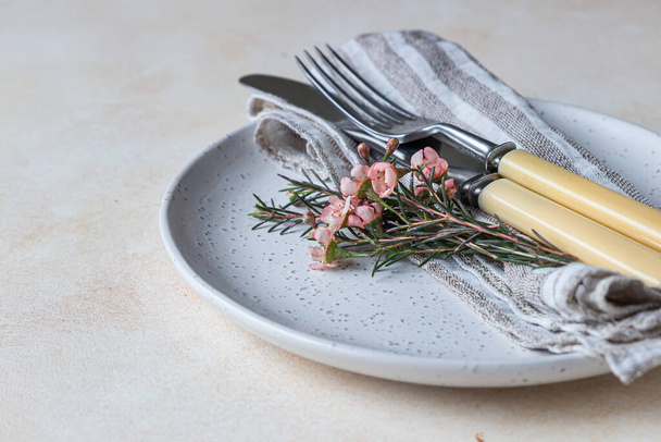 Romantic or spring table setting. Knife and fork, little pink flowers and linen napkin on a plate, light concrete background. - Foto, Bild